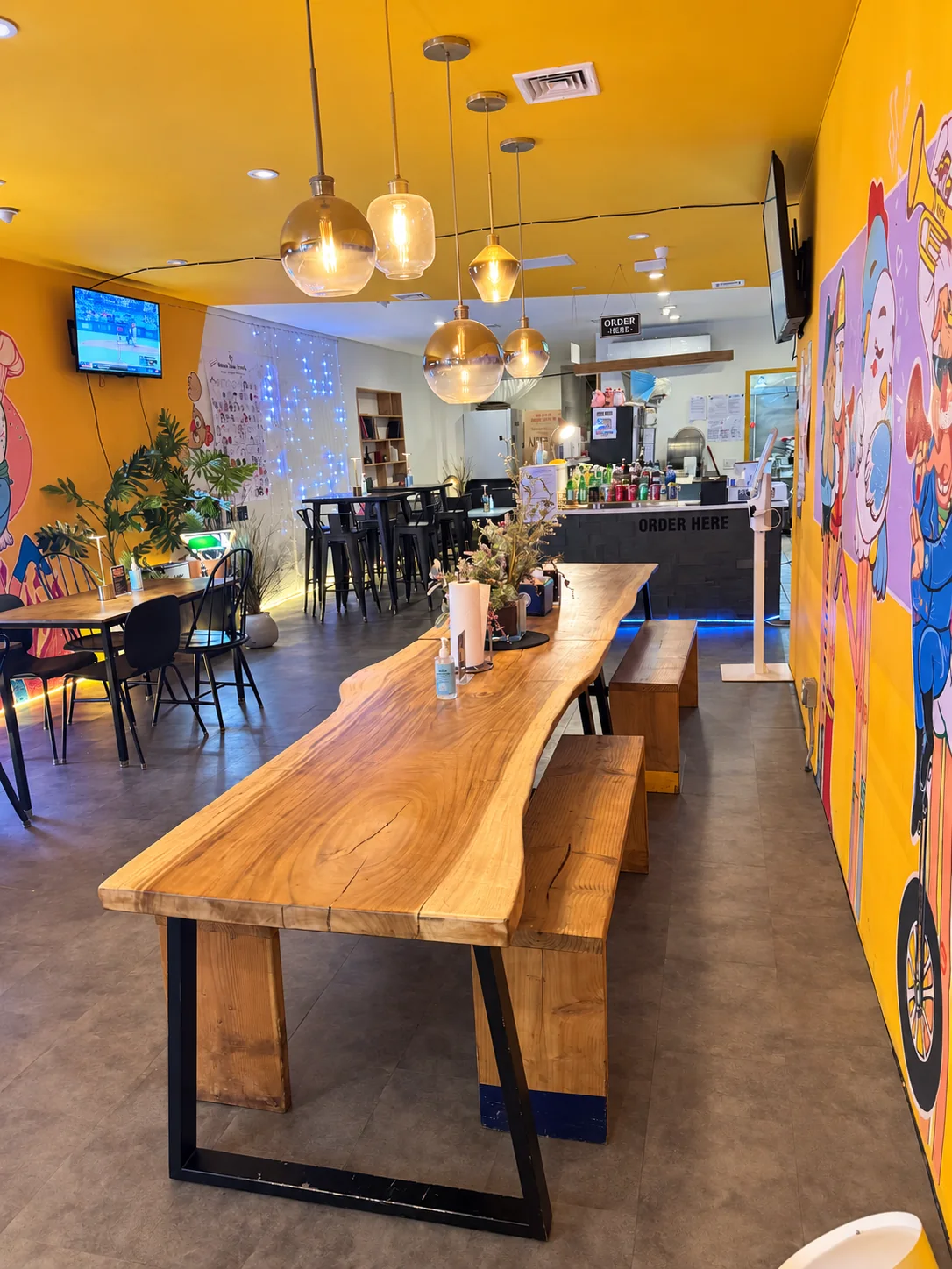 Inside Chicken Is Back: the long wooden table, pendant lights and yellow chicken murals leading to the order counter