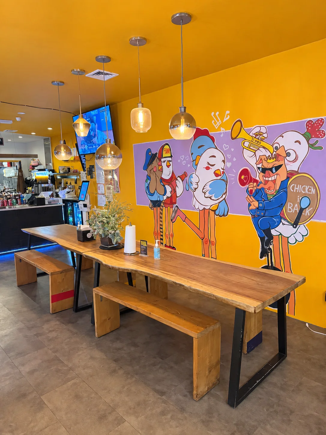 Chicken Is Back’s yellow dining room, with its chicken-band mural, glass pendant lights and long wooden table