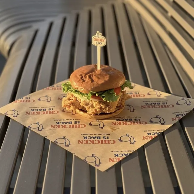 A thigh burger on Chicken Is Back paper, on a bench at sunset