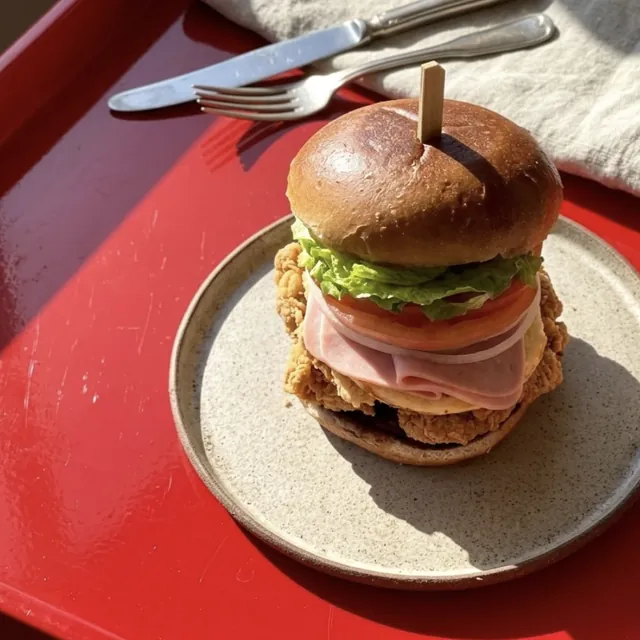 A thigh burger with ham and egg on a speckled plate, on a red tray in the sun